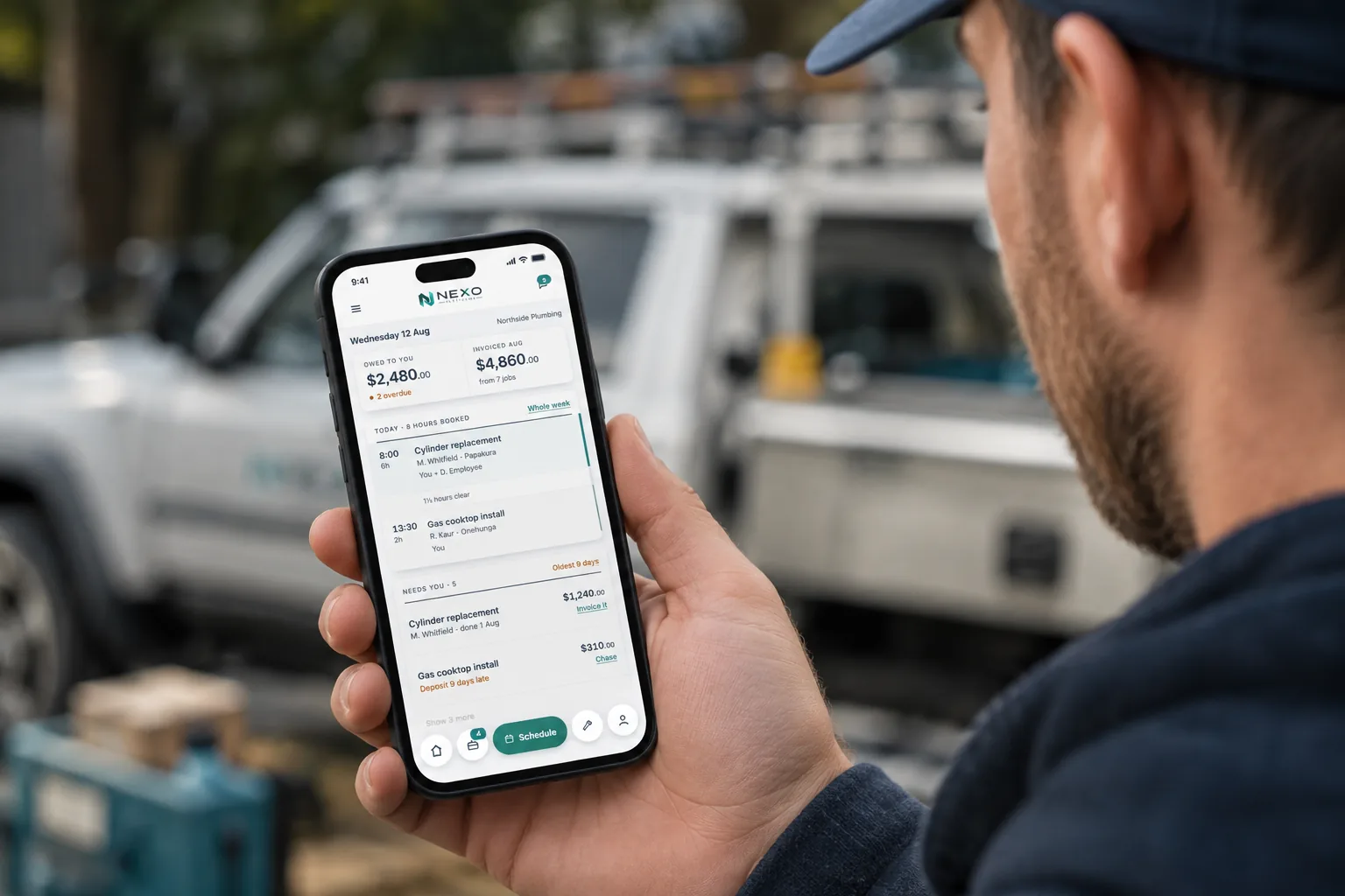 Someone in a work jacket and cap holds a phone showing the Nexo app, with a ute parked behind.